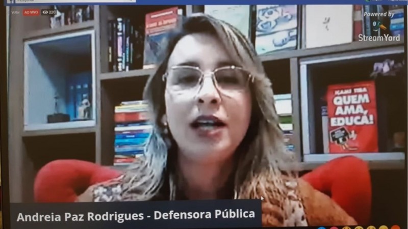 Defensora Pública Andreia Paz Rodrigues fazendo sua fala na live, com um livro vermelho ao fundo com o dizer: Quem Ama Educa!