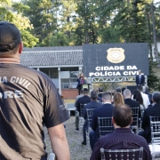 policial em primeiro plano e de costas, foco na sua camiseta com os dizeres "polícia civil core". ao fundo, a chefe de polícia discursando em cima do palco e, ao meio, o público acompanhando