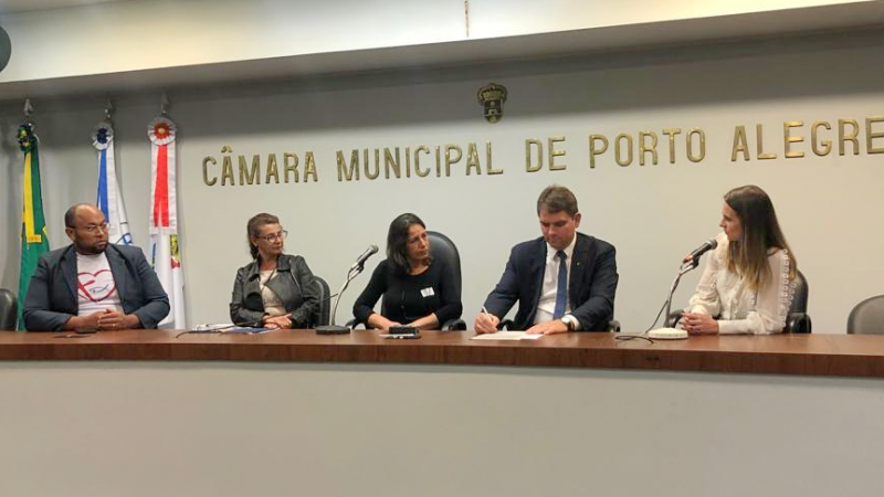 imagem da mesa da câmara, com letreiro atrás, os participantes sentados à mesa e a defensora pública falando