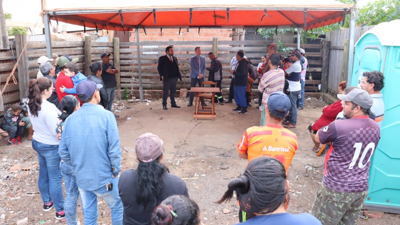 Foto dos catadores em círculo, em um terreno baldio com um gazebo laranja, escutando o defensor público falar