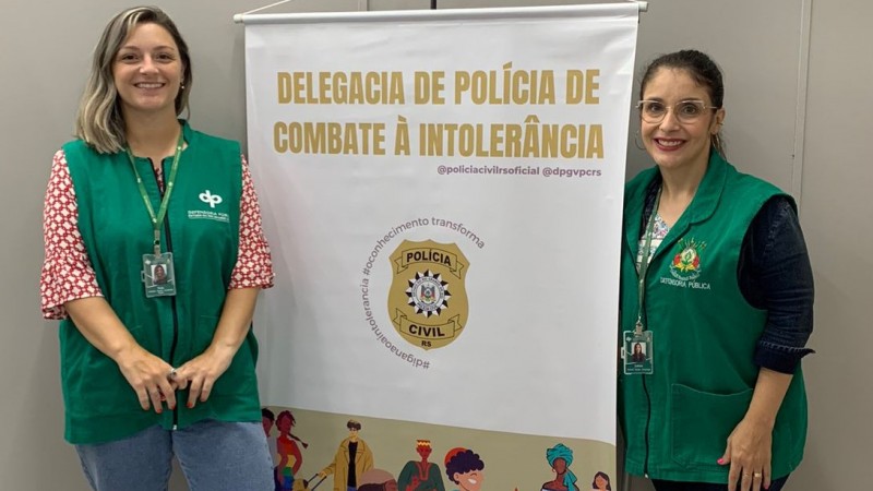 Thais Dalla Rosa e Leticia Souza Mello, entre elas o banner da Delegacia de Polícia de Combate à Intolerância