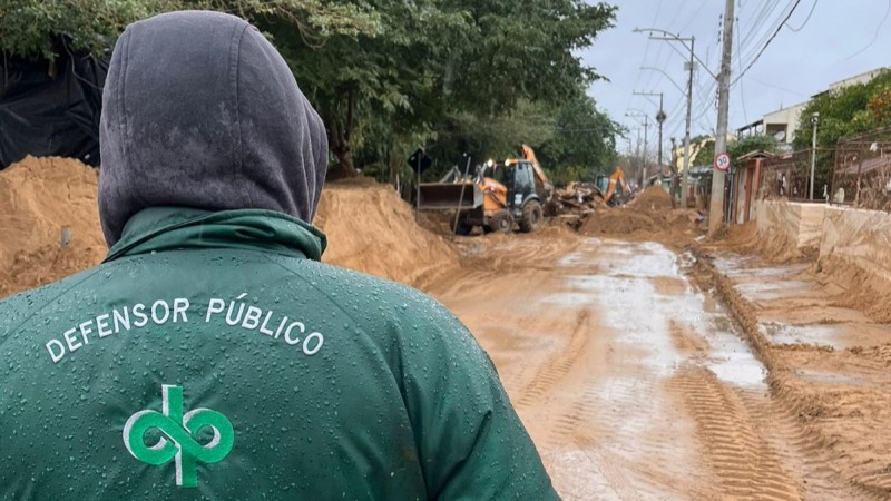 foto de um defensor público, de costas, olhando para a rua cheia de barro e água e, mais a frente, máquinas trabalhando para limpar o acesso