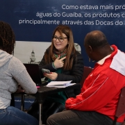 Na foto, há três pessoas sentadas em volta de uma mesa, duas delas estão de costas e a que está de frente está sorrindo enquanto conversa