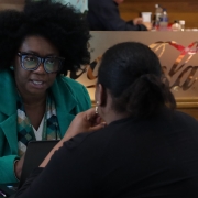 Na foto, há duas mulheres sentadas conversando, uma está de costas e a outra de frente