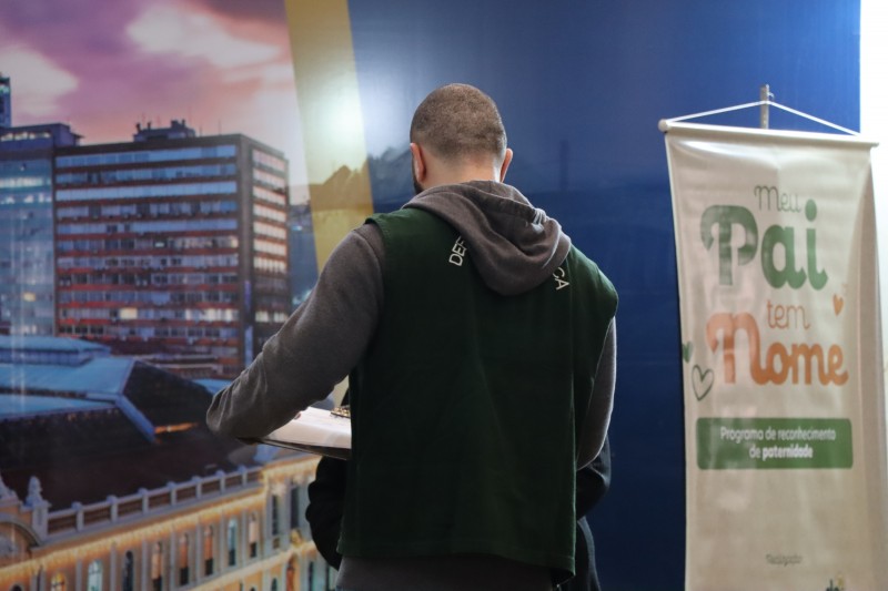 Homem está de costas, vestindo um colete verde em que se lê Defensoria Pública. Ao fundo, há um banner com os dizeres meu pai tem nome, programa de reconhecimento de paternidade