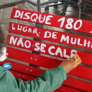 Na imagem vemos uma Defensora com pincel e tinta branca. Lê-se Disque 180.