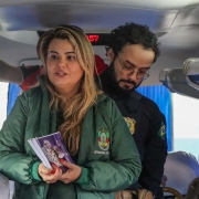 Em primeiro plano, defensora pública segura cartilhas de conscientização, em segundo plano há um policial federal. Estão dentro de um ônibus.
