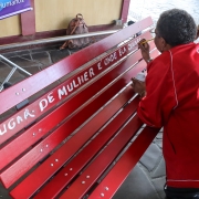 Mulher vestida de vermelho escreve em banco vermelho com pincel e tinta branca. Lê-se: lugar de mulher é onde ela quiser.