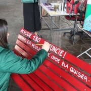 Mulher escreve no banco vermelho em tinta branca. Lê-se: Disque 180