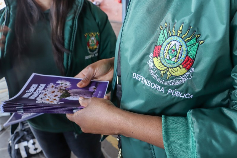 Mulher veste colete verde da defensoria pública e segura cartilhas. Lê-se: Violência Doméstica.