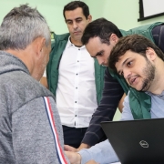 Em primeiro plano, um homem de costas escuta outro que conversa com ele. Este segundo está de frente e junto a um notebook. Ao fundo, há outros dois homens olhando para um objeto que está fora de cena.
