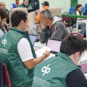 Três homens com coletes da Defensoria Pública, em primeiro plano, mexem em computadores tipo notebook. Um homem é atendido por um desses, e todos estão sentados. Ao fundo, é possível ver outras pessoas que aparentam estar recebendo atendimento.