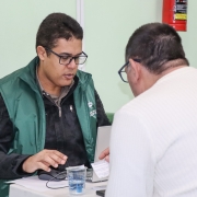 Em primeiro plano, um homem de costas segura um pequeno papel e está virado para outro homem que, de frente para a foto, manuseia um computador estilo notebook.