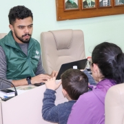 Uma mulher com uma criança, ambas de costas, estão viradas para um homem que, de frente, olha para a mulher com as mãos em um computador.