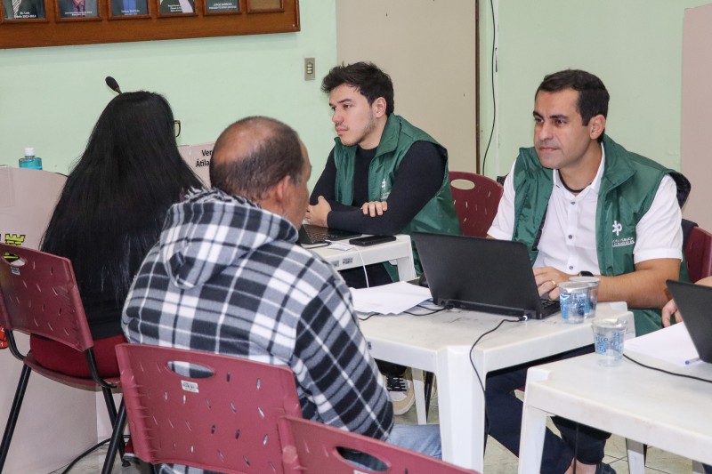 Uma mulher e um homem, sentados e de costas, estão de frente para dois homens, também sentados, mas de frente e com computadores em cima de mesas.