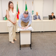 Homem assinando papel em pequena mesa ao lado de mulher que está de pé. Ao fundo, uma bancada com 4 pessoas.