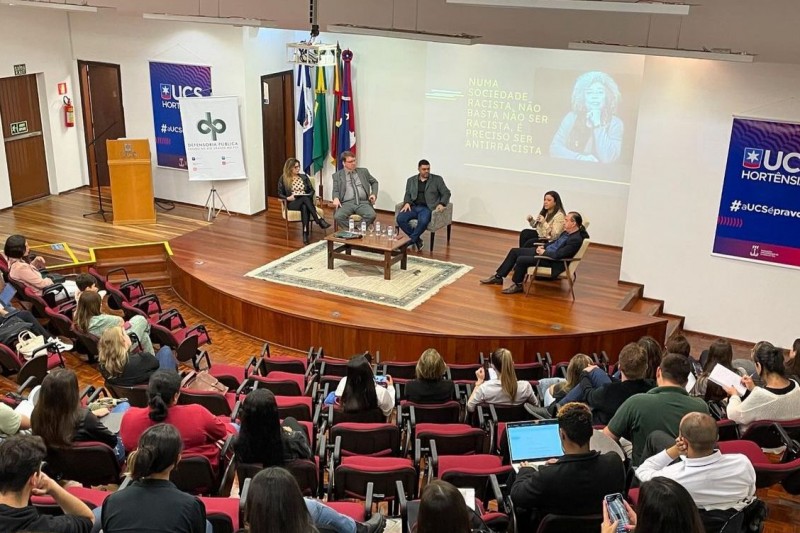 Vista superior de um auditório lotado, com um palco composto por 5 pessoas. Dentre elas, Alessandra Quines, defensora pública.