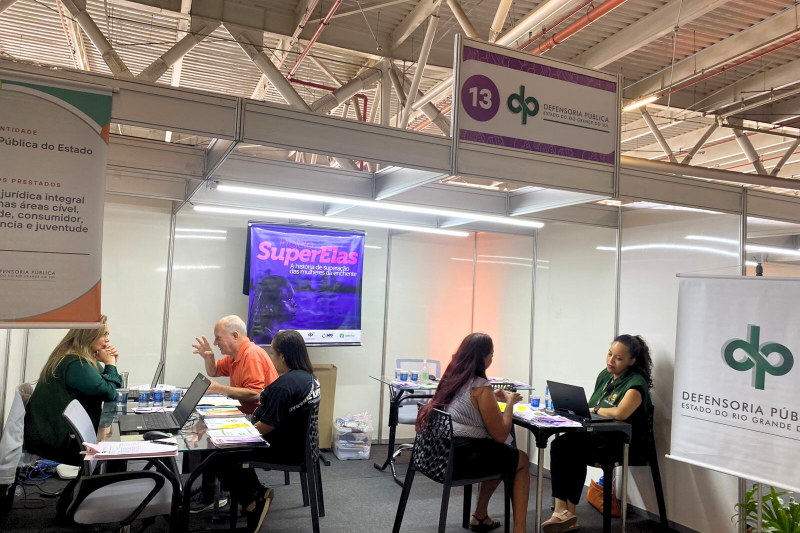 Duas defensoras p&uacute;blicas atendem tr&ecirc;s pessoas, l&ecirc;-se nos cartazes SuperElas (programa da Defensoria) e Defensoria P&uacute;blica