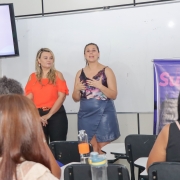 Duas mulheres estão de pé, uma ao lado da outra, falando para uma turma de mulheres adultas em uma sala de aula. 