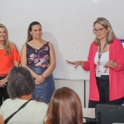 Uma mulher fala e gesticula em uma sala de aula para uma turma de mulheres adultas, sentadas nas cadeiras. Ao lado dela, duas outras mulheres também de pé olham para ela.