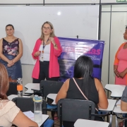 Uma mulher, de pé e de frente para a foto, fala em uma sala de aula para mulheres adultas sentadas em cadeiras de costas para a foto. Outras 3 mulheres estão ao lado da que está de pé falando.
