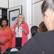 Em primeiro plano, de costas para a imagem, mulheres sentadas a mesas em uma sala de aula. Em segundo plano e de frente para a foto, uma mulher de pé fala com as alunas sentadas, e uma outra ao seu lado a observa.