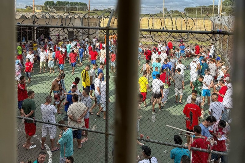Por trás de grades que aparecem desfocadas em um primeiro plano, ao fundo é possível ver um amontoado de homens andando e conversando em uma quadra,