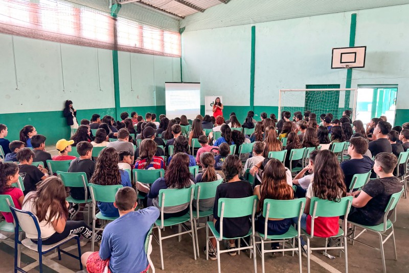 imagem da palestra sendo realizada em um ginásio, com a defensora de frente e dezenas de alunos de costas