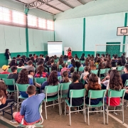 imagem da palestra sendo realizada em um ginásio, com a defensora de frente e dezenas de alunos de costas