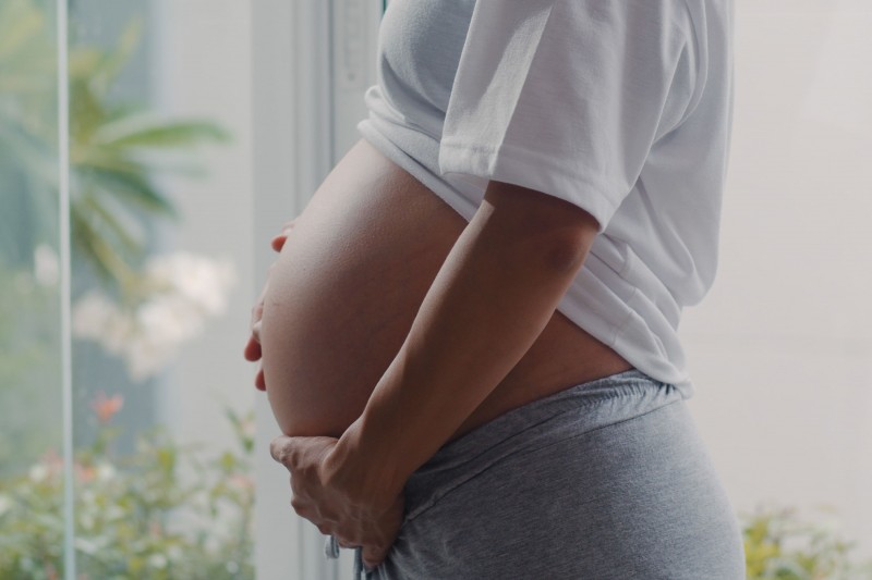 Mulher gr&aacute;vida com as m&atilde;os na barriga, vestindo blusa branca e cal&ccedil;a cinza.