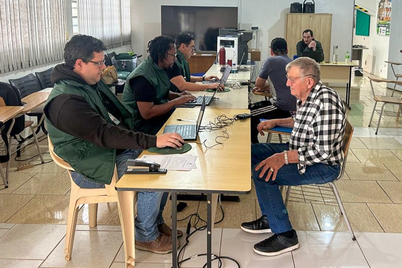 foto de uma sala com mesas e cadeiras, 4 servidores atendendo e 2 pessoas sendo atendidas