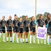 Equipe feminina do grêmio alinhada segurando a bandeira "Diga não à violência contra a mulher" na cor Lilás.