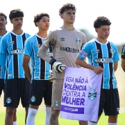 Categoria de base do Grêmio alinhada segurando a bandeira "Diga não à violência contra a mulher" na cor Lilás.