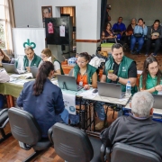 foto da equipe da defensoria,composta por 7 pessoas, em uma mesa comprida e de frente para a foto e duas mulheres e um homem sendo atendidos, de costas para a foto