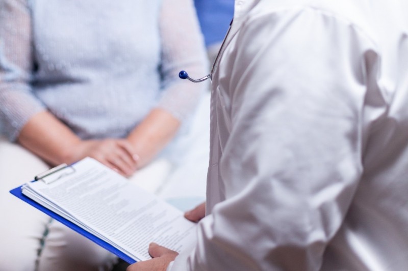Paciente e m&eacute;dico est&atilde;o sentados, frente a frente. O m&eacute;dico est&aacute; com uma planilha em que aparecem papeis escritos