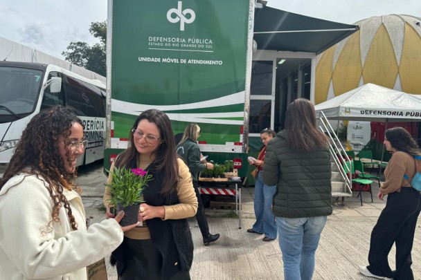 Foto mostra mulher recebendo flor das m&atilde;es de defensora p&uacute;blica e movimenta&ccedil;&atilde;o de servidoras da Defensoria P&uacute;blica em frente ao &ocirc;nibus da Defensoria no parque de exposi&ccedil;&otilde;es Assis Brasil.