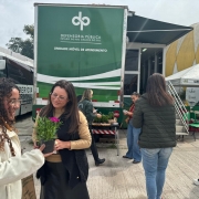 Foto mostra mulher recebendo flor das m&atilde;es de defensora p&uacute;blica e movimenta&ccedil;&atilde;o de servidoras da Defensoria P&uacute;blica em frente ao &ocirc;nibus da Defensoria no parque de exposi&ccedil;&otilde;es Assis Brasil.