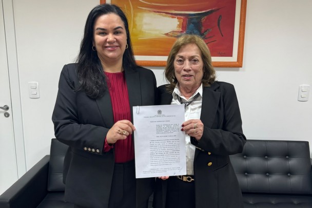 Mulher de cabelos escuros e vestindo blazer preta e camisa vermelha e mulher de cabelos claros e blazer escuro sorriem e seguram documento  