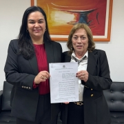 Mulher de cabelos escuros e vestindo blazer preta e camisa vermelha e mulher de cabelos claros e blazer escuro sorriem e seguram documento  